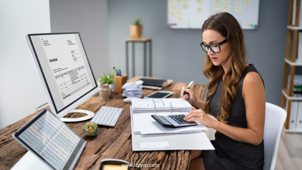 Woman using a calculator while reviewing invoices and financial documents at her desk, with accounting software open on dual monitors.